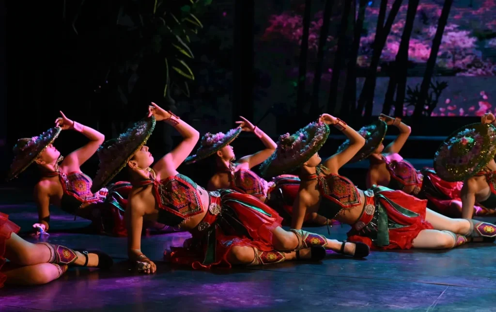 Miao ethnic minority dancers in embroidered red costumes and traditional flower-decorated hats performing a synchronized floor routine at the Charming Xiangxi cultural show, Zhangjiajie