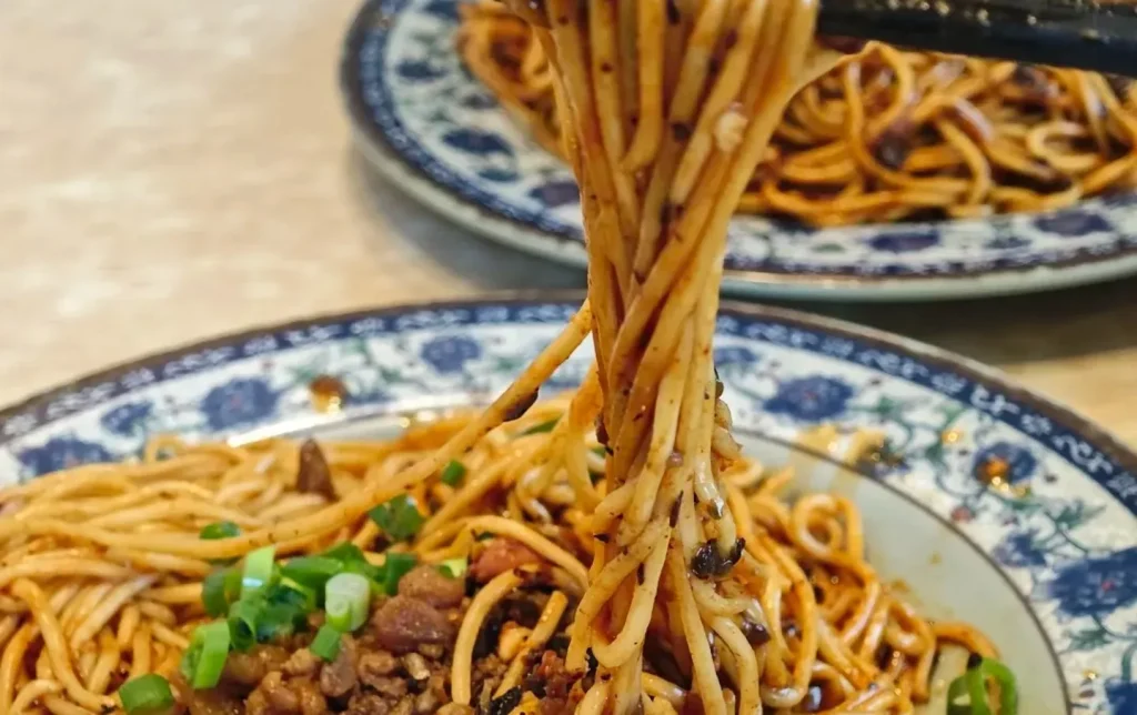 Chengdu dan dan noodles lifted by chopsticks with spicy minced pork sauce on traditional blue white plate