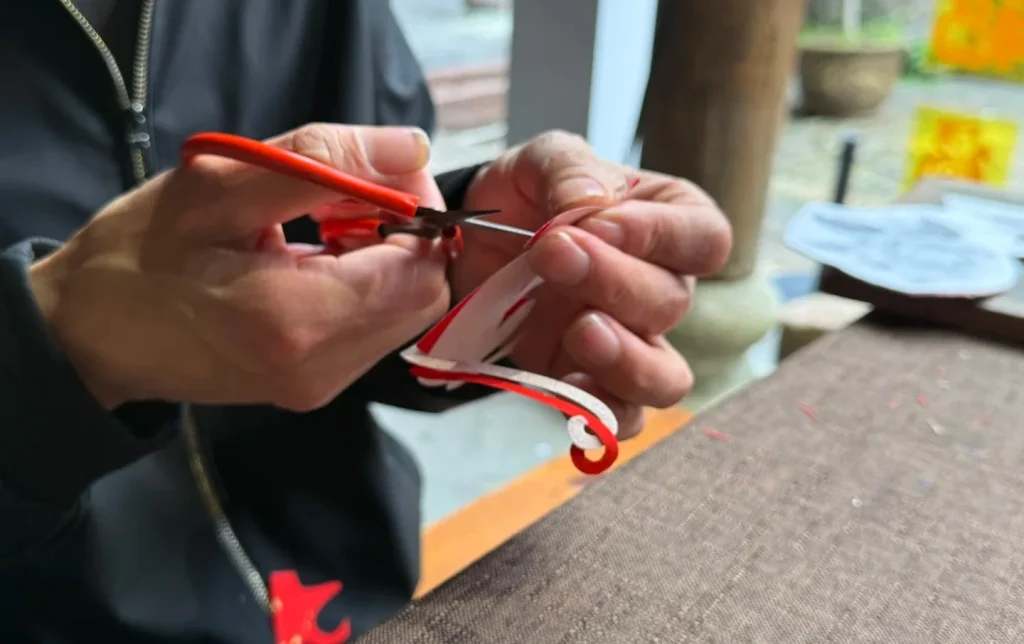 Artisan's hands using small red-handled scissors to make precise cuts along a curved red paper-cutting design at an outdoor workshop, showing the meticulous hand skill of Chinese jianzhi paper-cutting craft