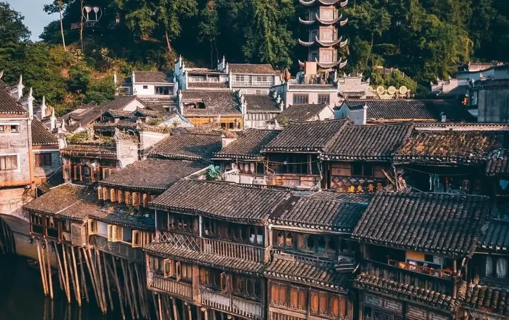 Traditional wooden stilted houses (diaojiaolou) of Fenghuang Ancient Town built on timber piles directly over the Tuojiang River, with a classical multi-tiered pagoda rising above the grey-tiled rooftops