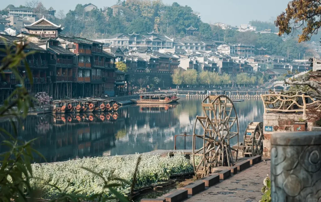 Traditional wooden water wheels and moored river boats reflected in the calm Tuojiang River at Fenghuang Ancient Town, with white wildflowers and centuries-old stilted wooden buildings lining the bank