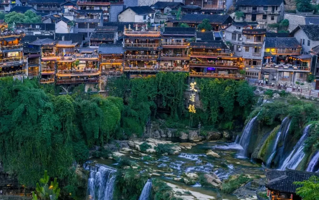 Furong Ancient Town (Furong Zhen) at blue-hour dusk with multi-tiered waterfalls tumbling through lush green cliff faces, terraced historic buildings glowing with warm lights on the hillside, Hunan