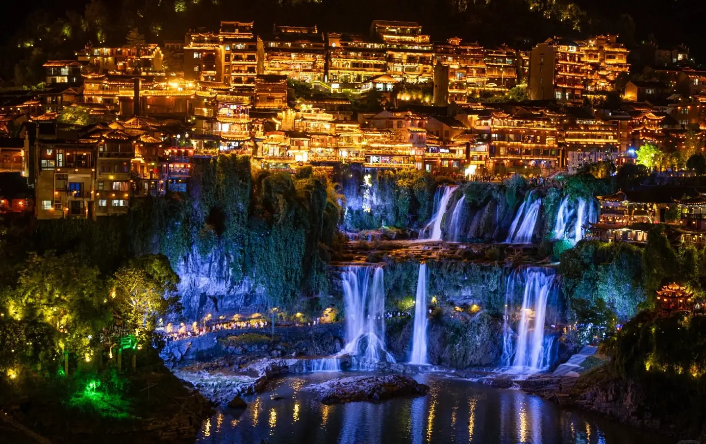 Furong Ancient Town at night with multiple blue-and-white illuminated waterfalls cascading down the cliffside while the historic hillside buildings glow in warm amber light above, Hunan, China