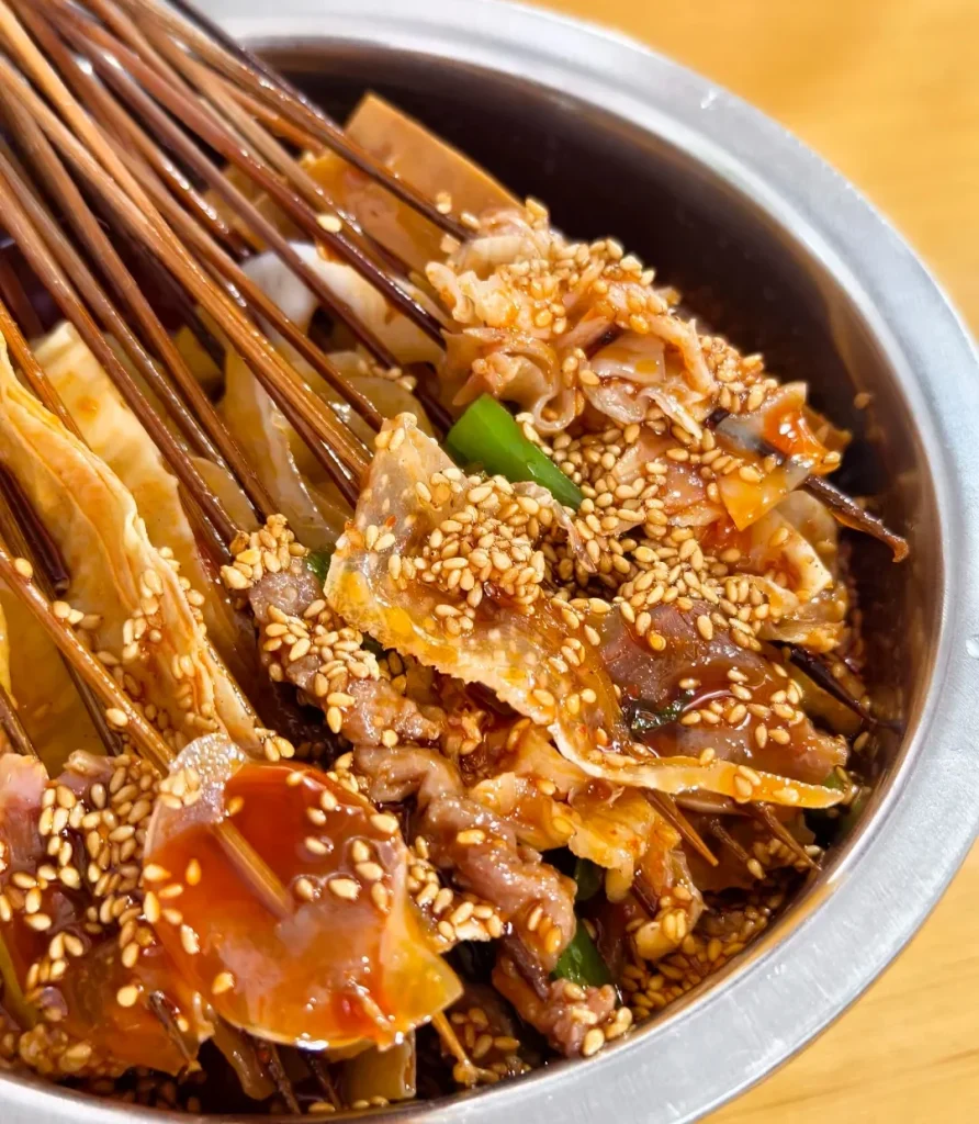 Leshan-style bobo chicken skewers with sesame sauce and mixed toppings in a metal bowl