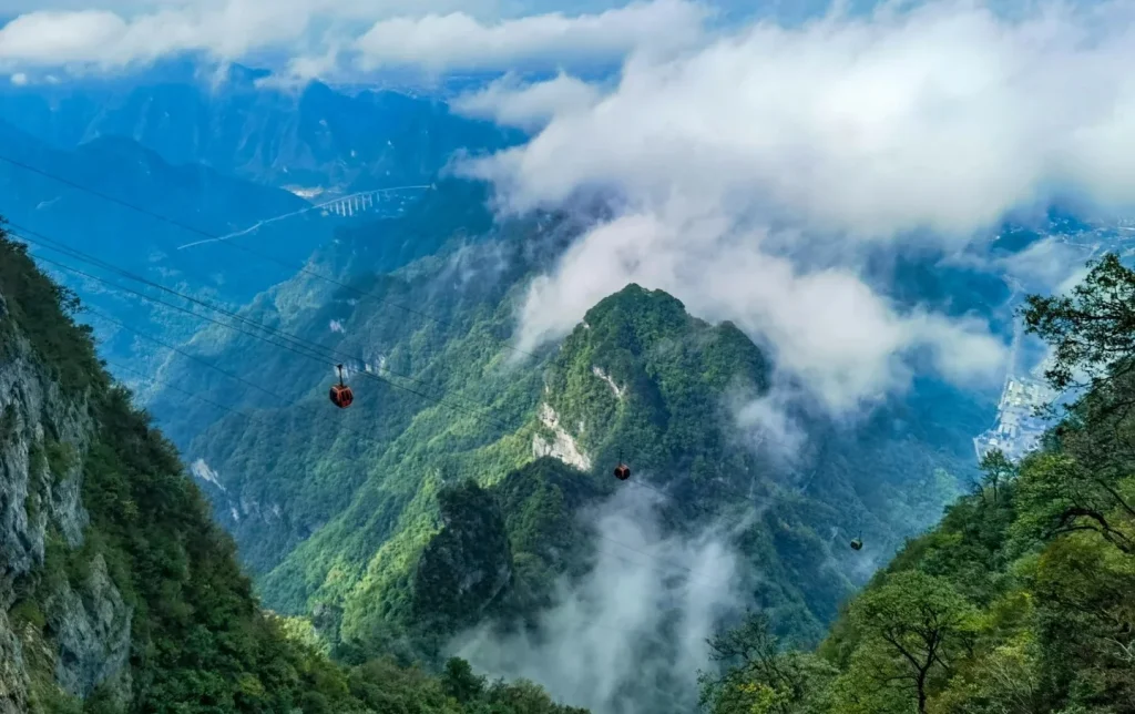 Red cable car gondolas of the Tianmen Mountain Ropeway gliding above a sea of clouds over lush green mountain peaks, with Zhangjiajie city visible far below in the misty valley