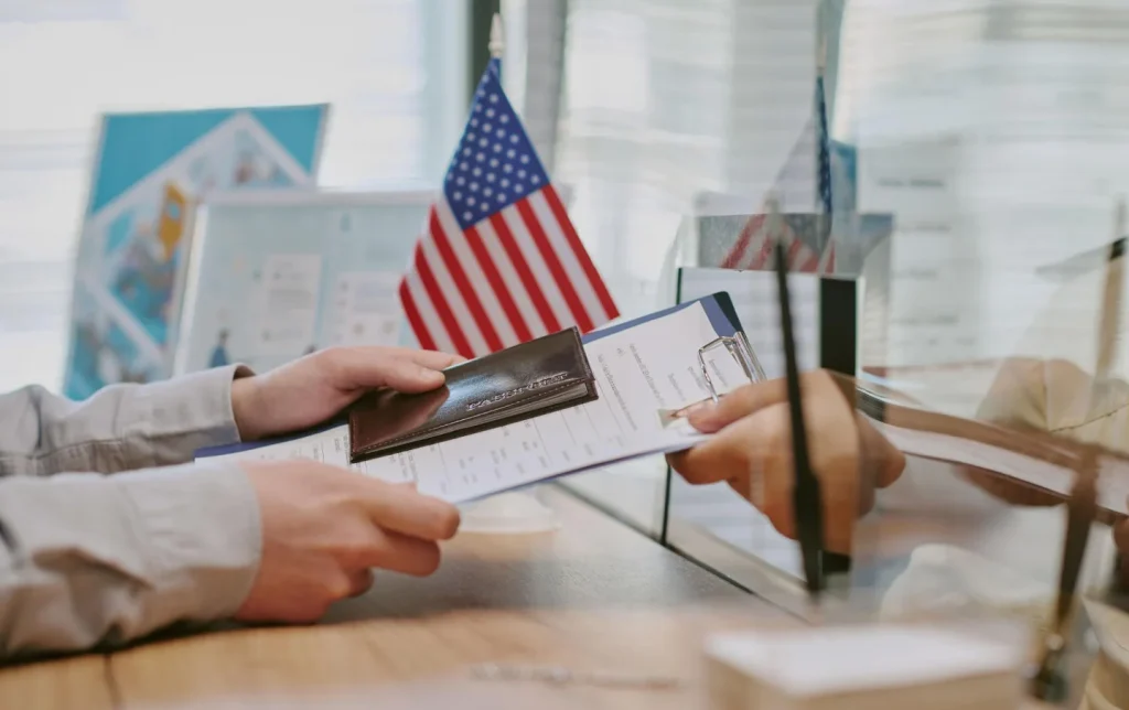 Traveler submitting passport and visa application form at a US immigration counter with an American flag in the background