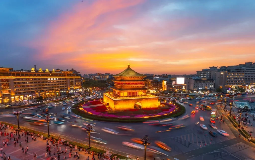 Long-exposure aerial view of Xi'an's Ming dynasty Bell Tower glowing in golden light at the center of a traffic roundabout beneath a vivid pink and blue sunset sky