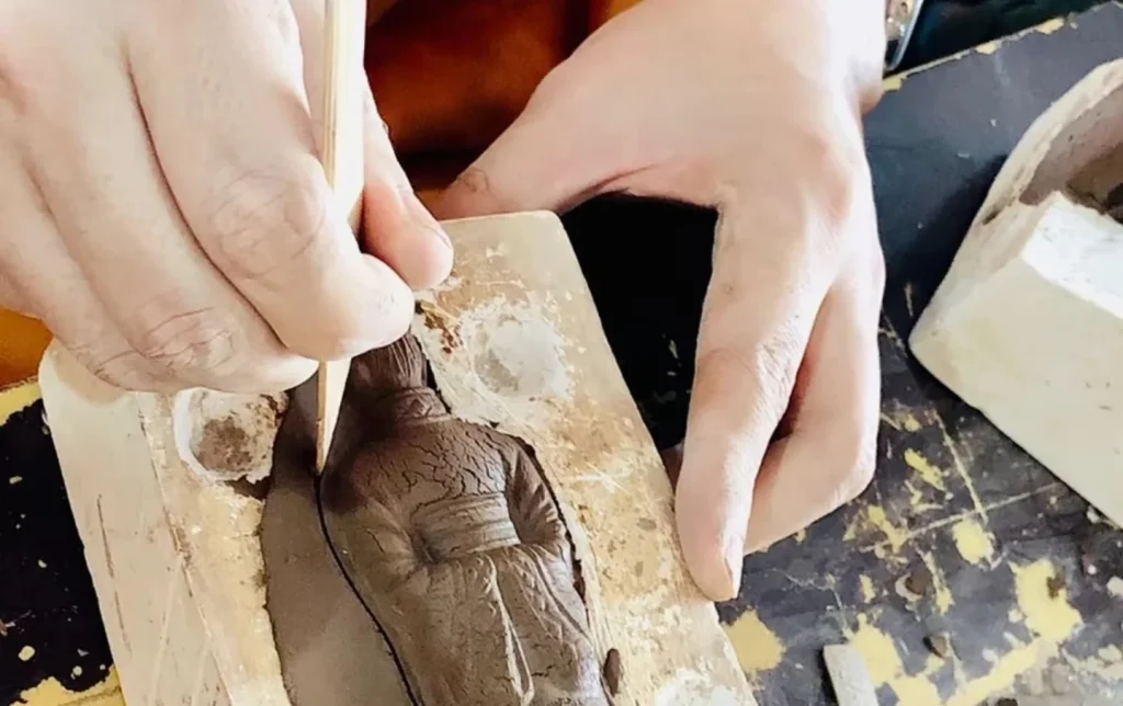 Hands using a wooden tool to carve fine details into a raw clay terracotta warrior figurine in a mold at a cultural heritage workshop in Xi'an