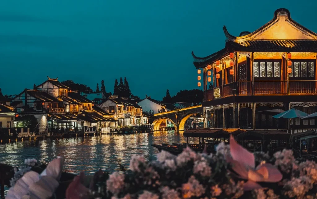 Glowing red lanterns reflecting on the canal of Zhujiajiao Water Town at night with a lit wooden pavilion and lotus blossoms in the foreground — Shanghai water town night trip