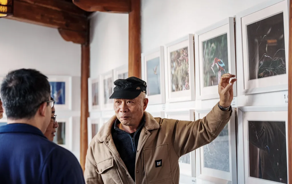 An elder explaining bird photography to visitors inside a traditional Chinese timber-framed gallery, an authentic cultural experience on China art and culture tours
